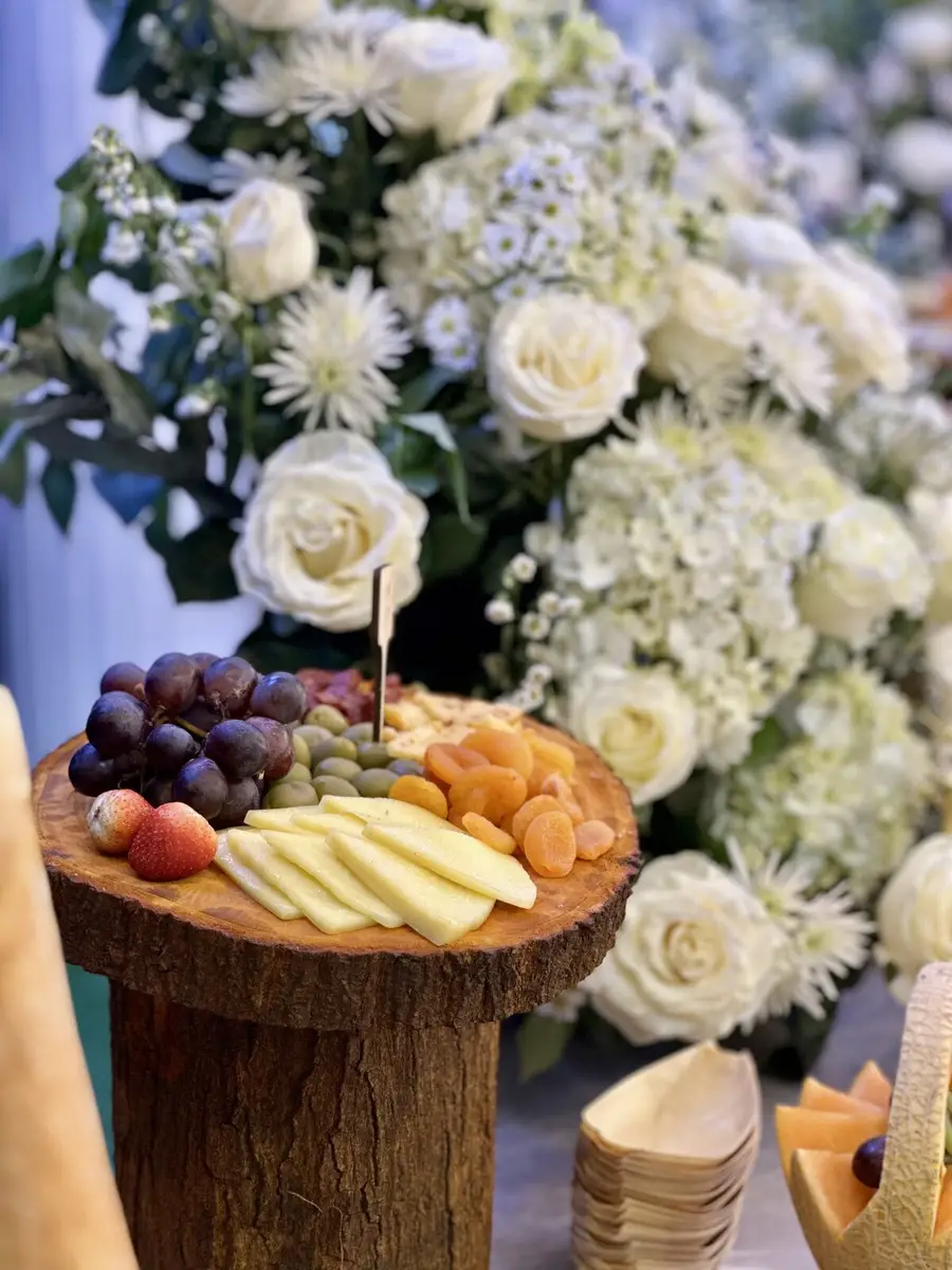 Tabla de quesos y frutas junto a un arreglo de flores blancas